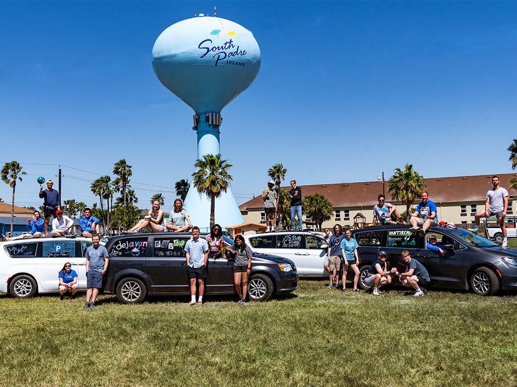 Group of people posing on and around vehicles in front of a water tower with "South Padre Island" written on it.